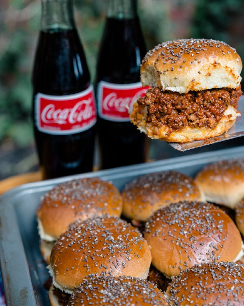 Homemade sloppy joes sliders with a bun that has seeds on top being served from a pan baked sliders with 2 coca-colas in the background as a serving suggestion.