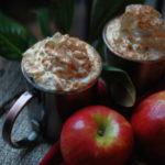 Two mugs of Caramel Apple Cider piled high with cinnamon whipped cream.