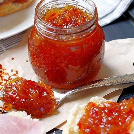 A jar of pineapple red pepper jelly with a spoonful of jelly and an open biscuit.