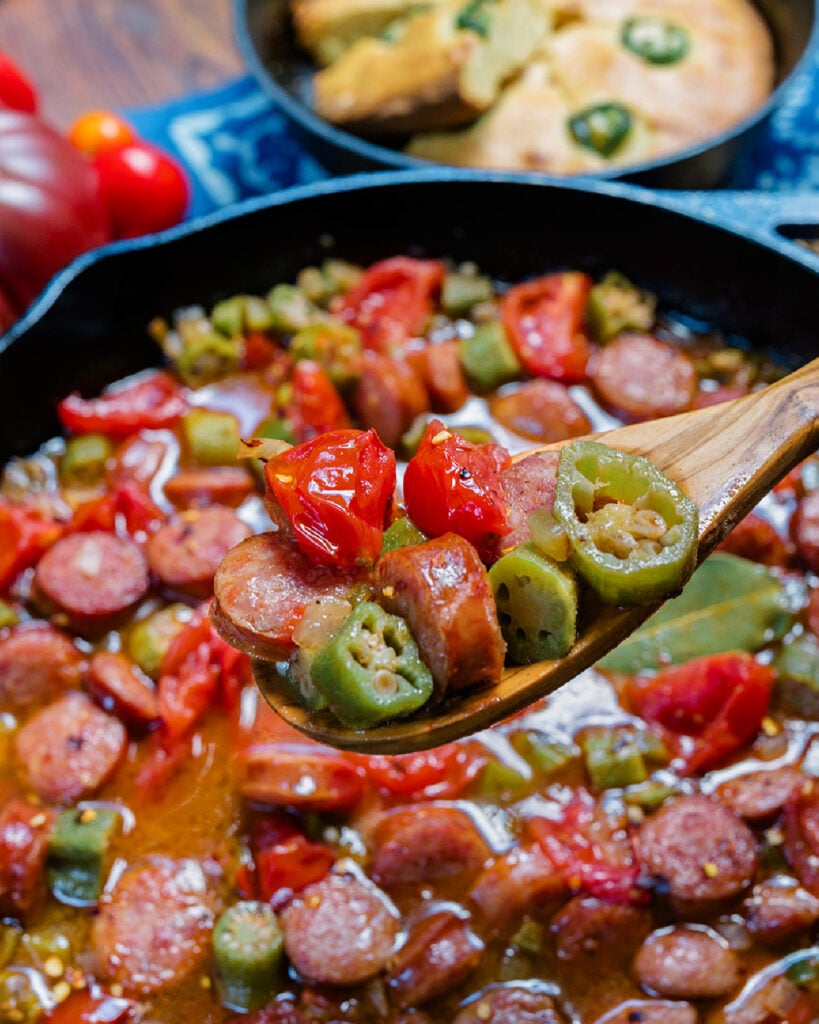 A spoonful of smoked sausage, tomatoes and okra on a wooden spoon after cooking showing proper color and texture, not overcooked or cooked down.