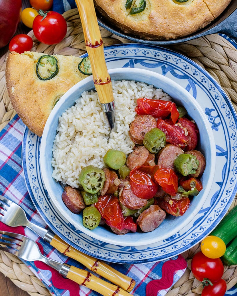 A bowl of stewed tomatoes and okra with smoked sausage served over rice with jalapeño cornbread on the side to show how this recipe can be enjoyed for dinner.