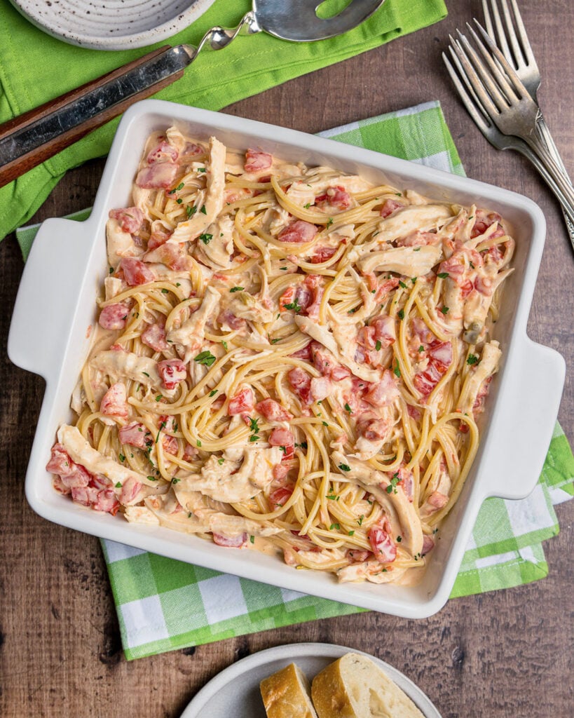 A baking dish filled with Rotel chicken spaghetti with green napkins and serving utensils.