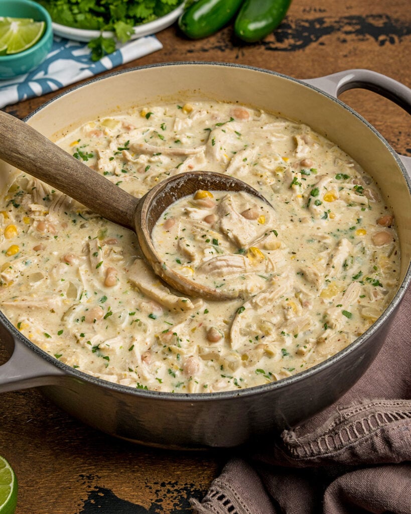 A dutch oven of warm chicken chili from the side with the serving ladle filled with chili to show the creamy thickness of the chili.