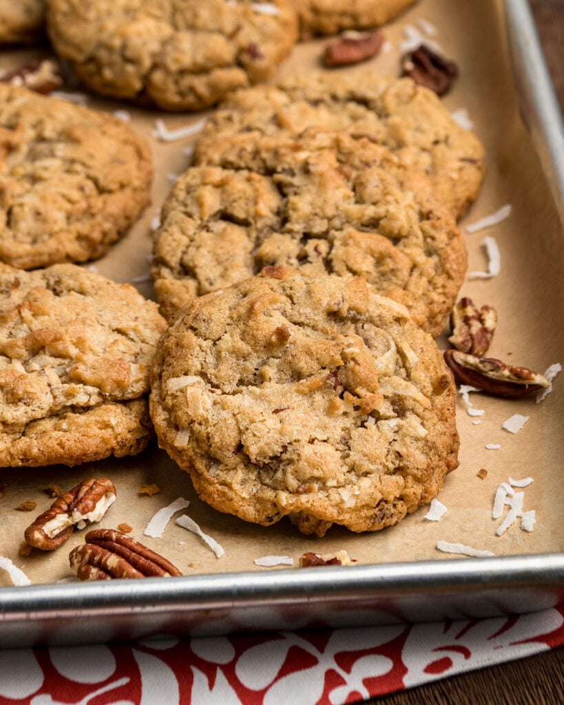 A close up of coconut pecan cookies on a baking sheet with parchment paper and extra coconut and pecans to show cookie texture and golden color.