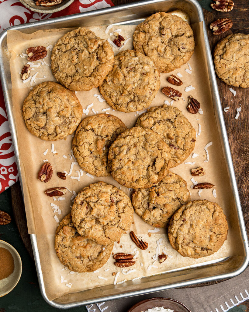 A pan lined with parchment paper and filled with pecan coconut cookies, extra pecans and coconut to show a dozen cookies on the baking sheet.