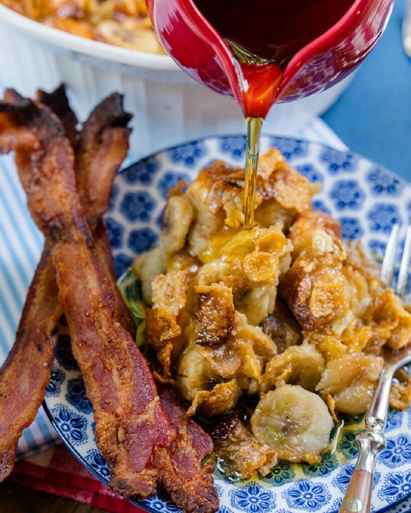 Maple syrup being drizzled over a serving of this french toast casserole with bananas with a side of bacon.
