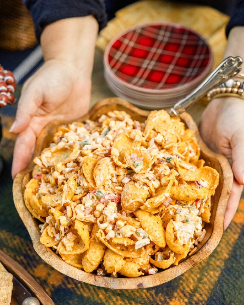 A wooden bowl filled with large Frtio corn chips, corn, green and red bell pepper, purple onion and a spicy creamy dressing to make a tailgate or cookout salad with lots of texture.