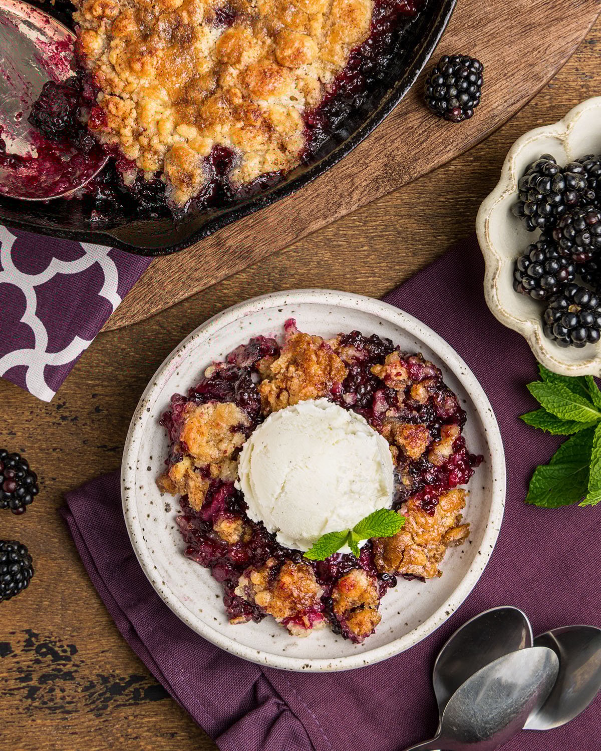 Looking down on a bowl of blackberry cobbler with a scoop of ice cream, mint garnish and skillet of cobbler in the background,