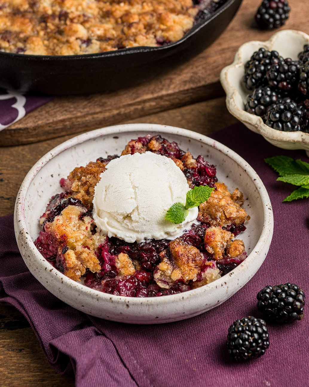 A bowl of blackberry cobbler with a crisp crust served with vanilla ice cream and a sprig of mint as an easy serving idea.