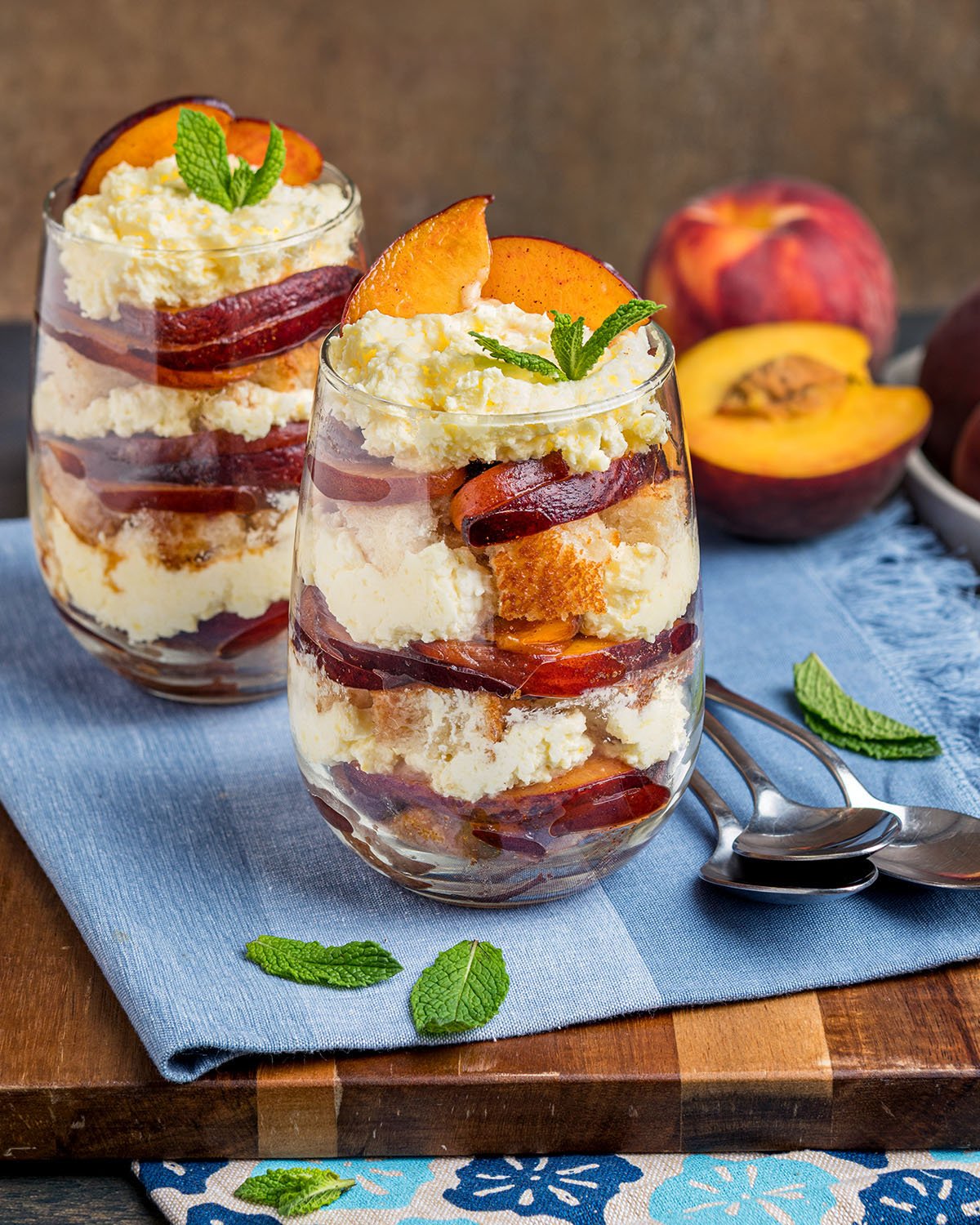 Two old fashioned peach trifles made in cups and surrounded by peaches to show this recipe is made with fresh fruit not canned peaches.