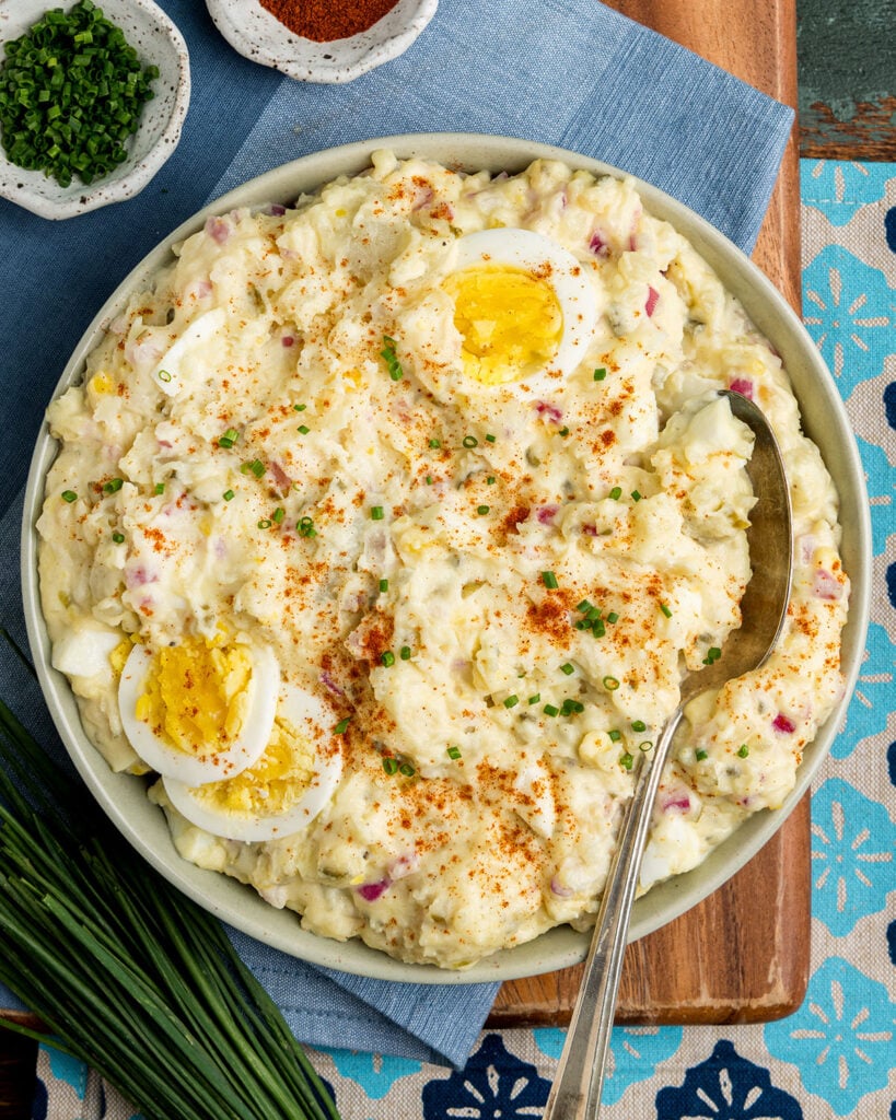 A serving bowl filled with yellow potato salad, topped with paprika, chopped chives and sliced boiled eggs as an eye catching serving idea