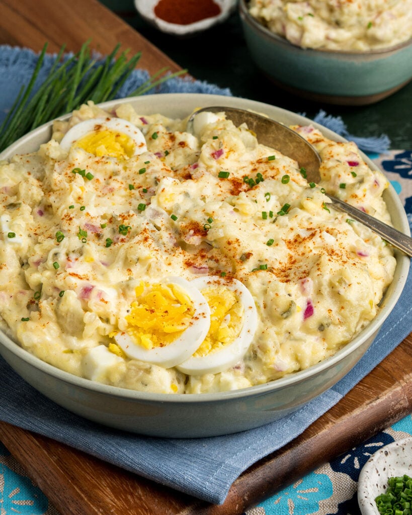 A serving bowl filled with traditional southern potato salad, chives, serving spoon and a blue dinner napkin as a cookout side dish idea.