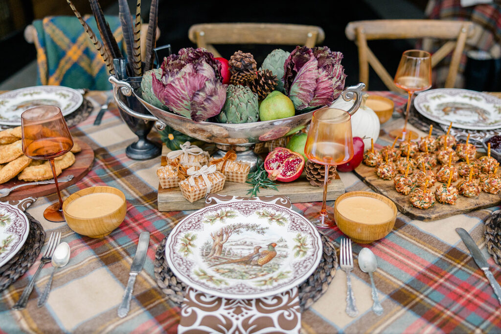 Spode Woodland place setting with pheasants, brown napkin and woven placemats on a plaid tablecloth with vegetables, fruit and pinecones in silver urn as a centerpiece.