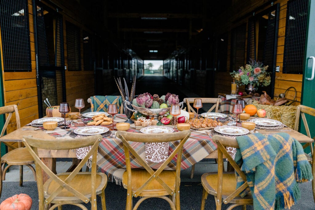 A rustic, cozy Friendsgiving table done in a plaids, wood & silver serving pieces Ralph Lauren style in front of a horse barn.